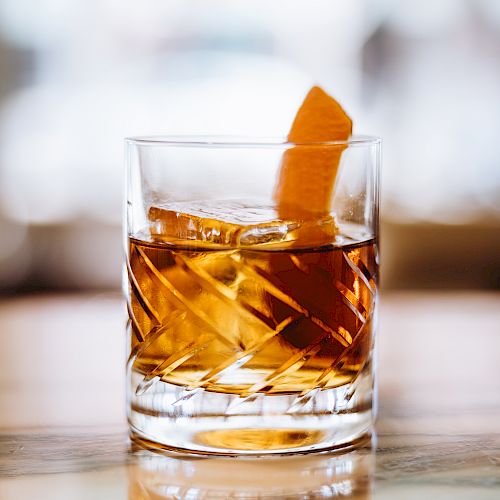 A glass of amber-colored liquid with ice and an orange peel garnish rests on a polished surface.