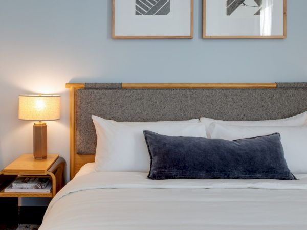 A neatly made bed with white linens and a gray pillow, a nightstand with a lamp, art on the wall, and a warm, inviting ambiance.
