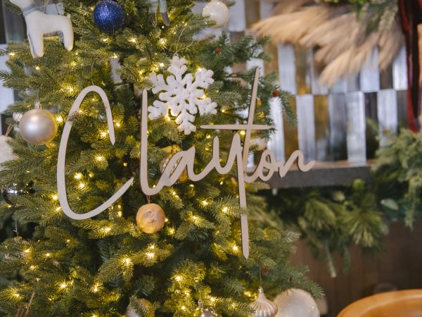 A Christmas tree with lights and ornaments, plus a wooden script name sign reading &ldquo;Clayton&rdquo; hanging among the branches.