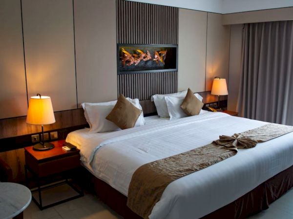 The image shows a neatly arranged hotel room with a large bed, pillows, bedside tables, lamps, and curtains, creating a cozy atmosphere.
