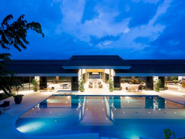 The image shows a modern building with a lit swimming pool under a blue sky at dusk, surrounded by trees and outdoor seating areas.