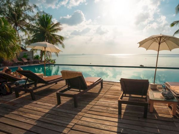 Lounge chairs and umbrellas on a wooden deck by a tranquil pool with a palm tree and ocean in the background under a bright sky.