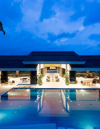 The image shows a luxurious building with a large, illuminated swimming pool in front, set against a twilight sky.