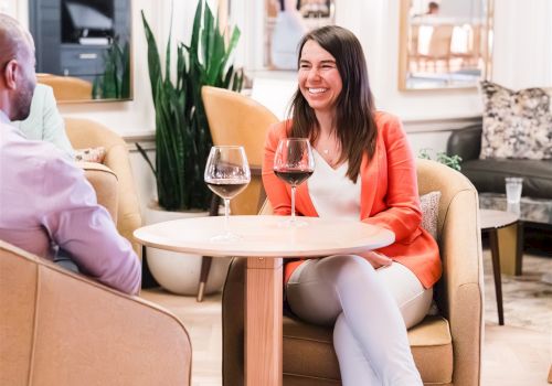 A woman smiling at a cozy cafe table with wine glasses, chatting with a man across from her in a bright, stylish lounge setting.