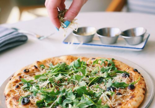 A hand sprinkles toppings onto a fresh arugula pizza with olives and cheese, while a tiny herb garden vibe fills the scene.