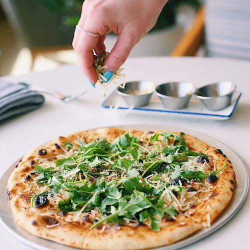 A hand sprinkles toppings onto a fresh arugula pizza with olives and cheese, while a tiny herb garden vibe fills the scene.