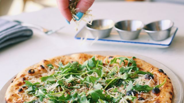 A hand sprinkles toppings onto a fresh arugula pizza with olives and cheese, while a tiny herb garden vibe fills the scene.