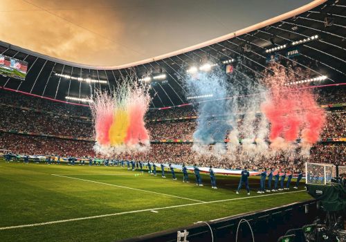 Colorful stadium fireworks explode as teams line up on a lush football pitch under bright stadium lights.