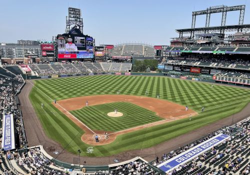 A sunny baseball stadium with a neatly mowed diamond, surrounding stands filled with fans, and a large video board at left.