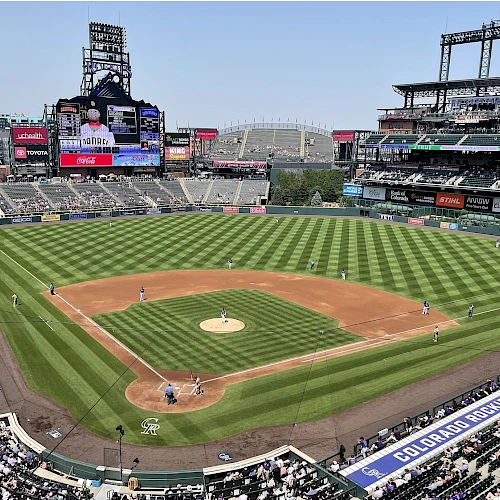 A sunny baseball stadium with a neatly mowed diamond, surrounding stands filled with fans, and a large video board at left.