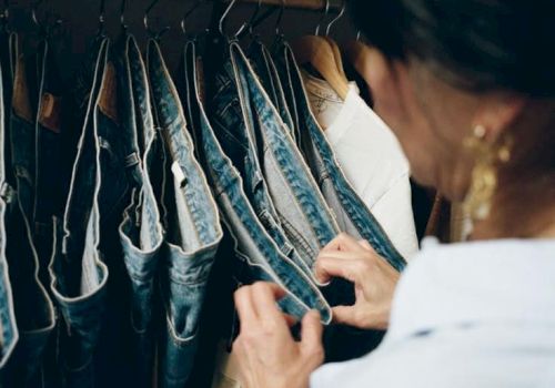 A person browses or arranges denim jeans on a rack, focusing on a pair held by the hands inside a closet. It&rsquo;s a casual shopping or organizing moment.