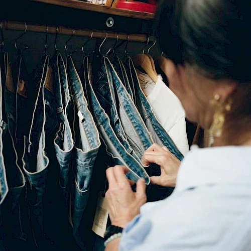 A person browses or arranges denim jeans on a rack, focusing on a pair held by the hands inside a closet. It&rsquo;s a casual shopping or organizing moment.