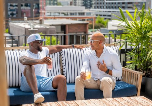 Two friends relax on a rooftop balcony, sipping drinks and chatting on a sunny day with city buildings in the background.