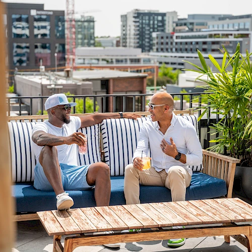 Two friends relax on a rooftop balcony, sipping drinks and chatting on a sunny day with city buildings in the background.