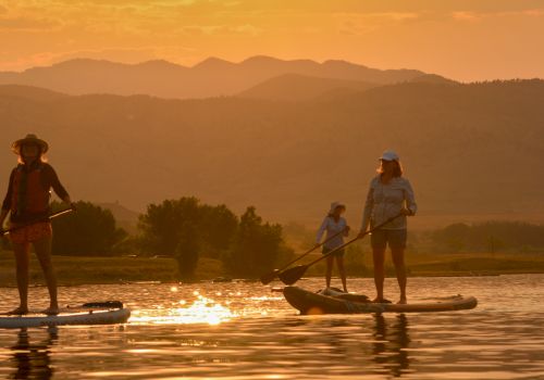 Three people stand on stand-up paddleboards at sunset, paddling across calm water with a golden mountain backdrop.