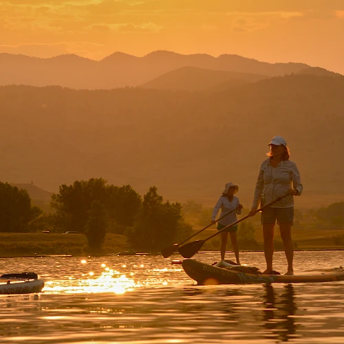 Three people stand on stand-up paddleboards at sunset, paddling across calm water with a golden mountain backdrop.