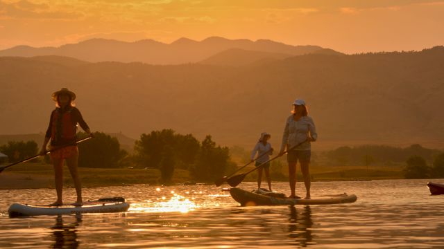 Three people stand on stand-up paddleboards at sunset, paddling across calm water with a golden mountain backdrop.