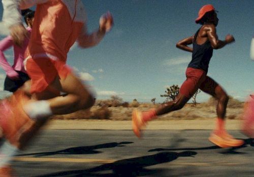 Runners sprint down a desert road, blurred in motion, wearing bright athletic gear and orange shoes under a clear, sunny sky.