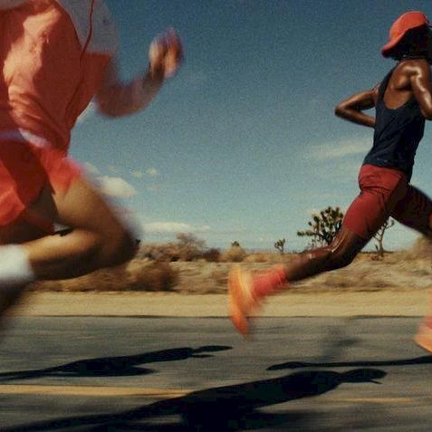 Runners sprint down a desert road, blurred in motion, wearing bright athletic gear and orange shoes under a clear, sunny sky.