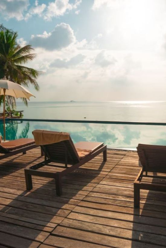 A wooden deck with sun loungers and umbrellas overlooks a serene infinity pool and ocean, framed by palm trees and a bright sky.