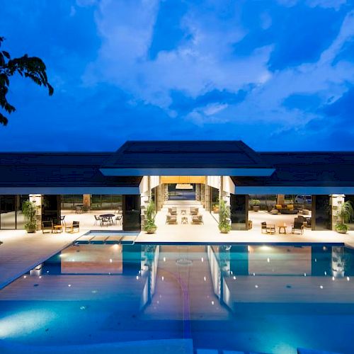 A luxurious poolside setting at dusk, featuring a modern building with illuminated interiors and seating areas, under a vibrant evening sky.
