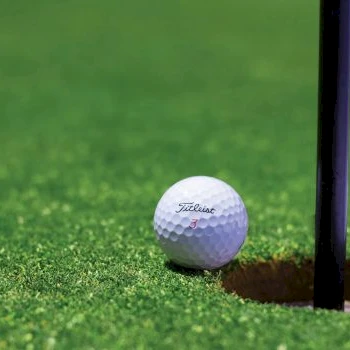 A golf ball sits near the hole on a green putting surface, close to the flagstick, ready for a potential putt.