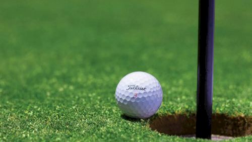 A golf ball sits near the hole on a green putting surface, close to the flagstick, ready for a potential putt.