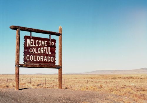 A weathered wooden sign reads &ldquo;Welcome to Colorful Colorado&rdquo; in a vast, arid desert landscape with a clear blue sky.