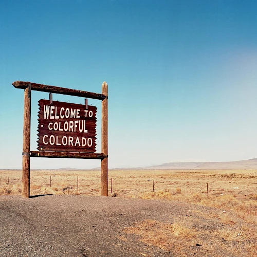 A weathered wooden sign reads &ldquo;Welcome to Colorful Colorado&rdquo; in a vast, arid desert landscape with a clear blue sky.