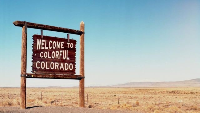A weathered wooden sign reads &ldquo;Welcome to Colorful Colorado&rdquo; in a vast, arid desert landscape with a clear blue sky.