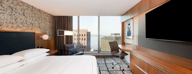 The image shows a modern hotel room with a bed, large TV, desk, chair, and a window with a city view, featuring contemporary decor.