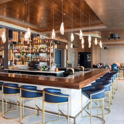 A modern bar with a wooden counter, blue stools, decorative lighting, and well-stocked shelves in a spacious setting.