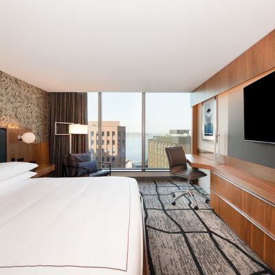 A modern hotel room features a bed, desk, large TV, and floor-to-ceiling window with city views, combining comfort and style.