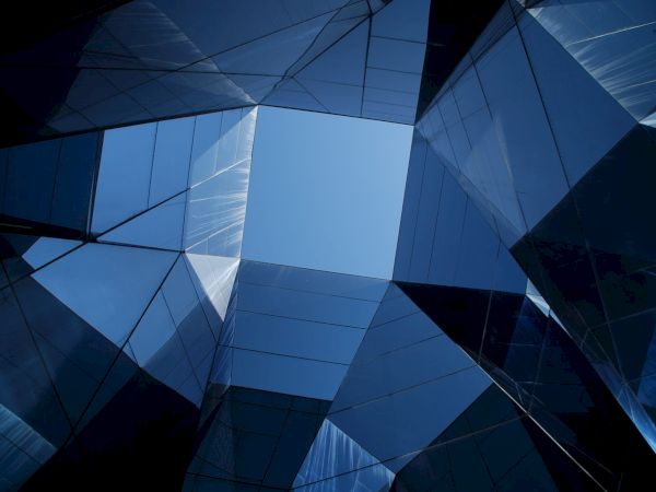 Geometric glass structures form a pattern, reflecting a blue sky above. The design creates an abstract, modern architectural view.
