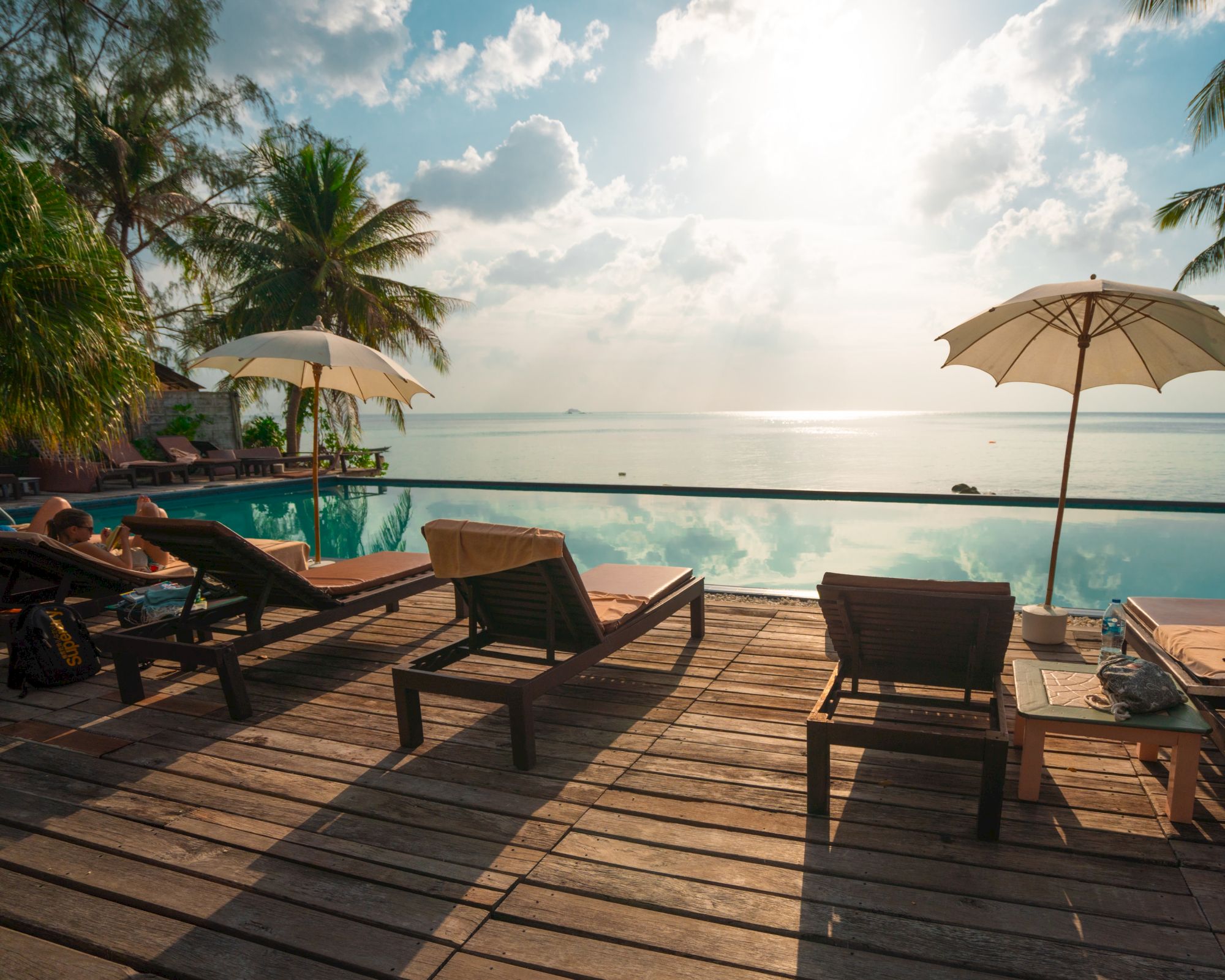 A serene poolside scene with lounge chairs, umbrellas, palm trees, and a view of the ocean under a bright sky.