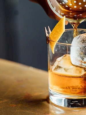 A cocktail being poured into a glass with ice and a citrus peel garnish on a bar counter.