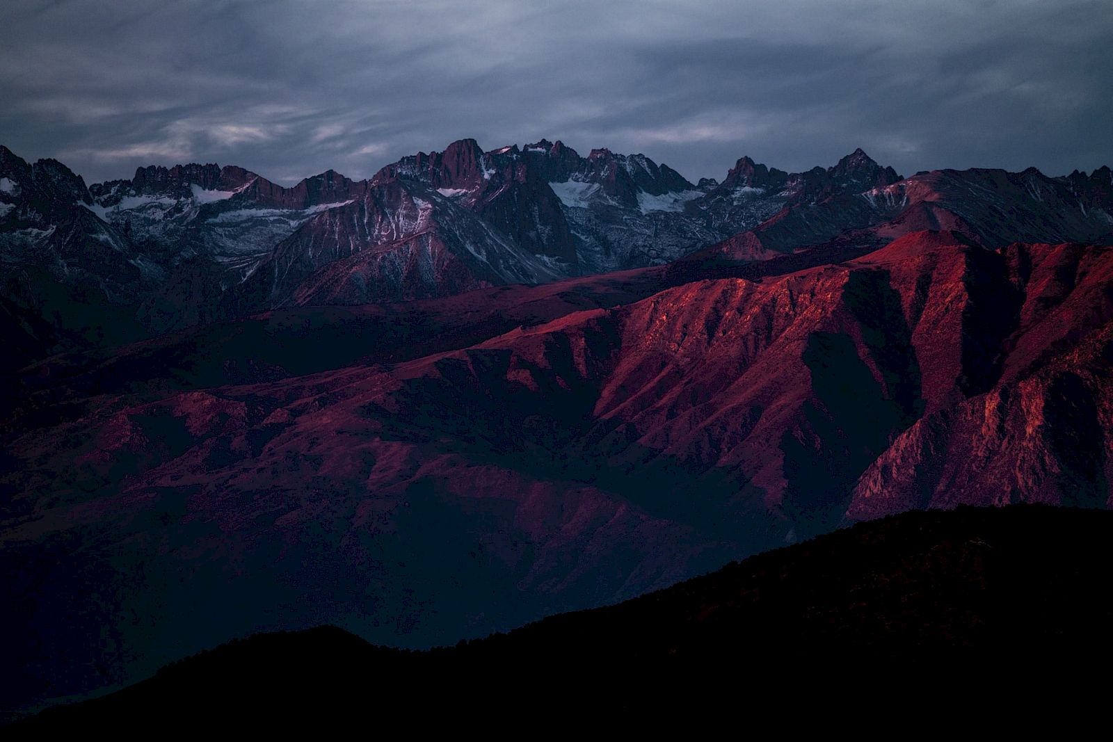 A mountain range under a dramatic sky, highlighted with deep red and blue hues at sunset, creating a striking and atmospheric scene.