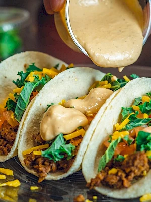 Three tacos filled with ground meat, cheese, and greens are being drizzled with sauce, served on a plate.
