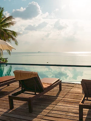 A serene beachside pool area with lounge chairs, umbrellas, and a view of the ocean under a partly cloudy sky.