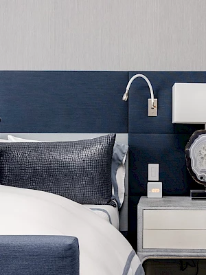 A modern bedroom features a bed with blue and white linens, a navy headboard, and a nightstand with a lamp and decorative item.