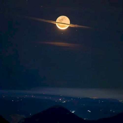A moon shines brightly above an illuminated landscape, framed by a horizontal cloud resembling Saturn's rings in a dark, serene night sky.
