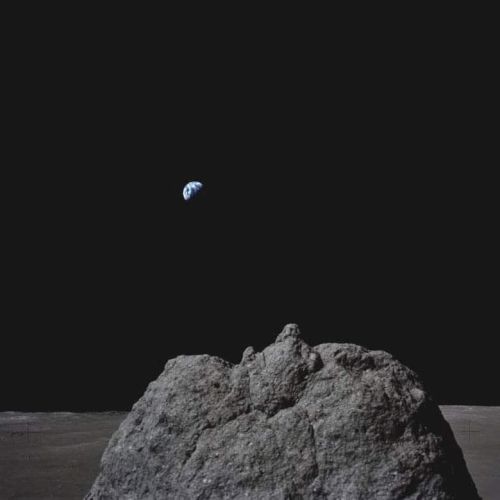 A view of Earth rising above the lunar horizon with a rocky surface in the foreground and a black sky in the background.