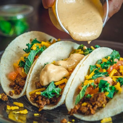 Three tacos on a plate with ground meat, lettuce, cheese, and sauce being poured over them, creating a tasty and colorful presentation.