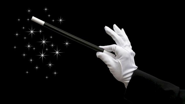 A gloved hand wields a wand over sparkling magic lights against a black background.