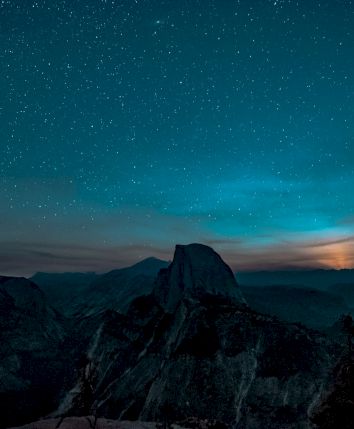 A starry night sky over a mountain landscape with a glowing horizon and silhouetted trees in the foreground.