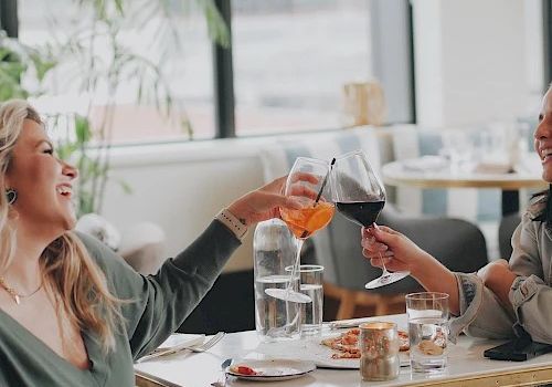 Two people clink glasses over drinks in a caf&eacute;, laughing as they toast together.
