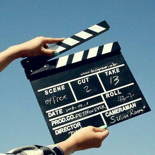 A person holds a black clapperboard movie slate with &ldquo;Scene,&rdquo; &ldquo;Take,&rdquo; and &ldquo;Roll&rdquo; markings against a blue sky, ending a filming moment.
