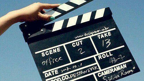 A person holds a black clapperboard movie slate with &ldquo;Scene,&rdquo; &ldquo;Take,&rdquo; and &ldquo;Roll&rdquo; markings against a blue sky, ending a filming moment.
