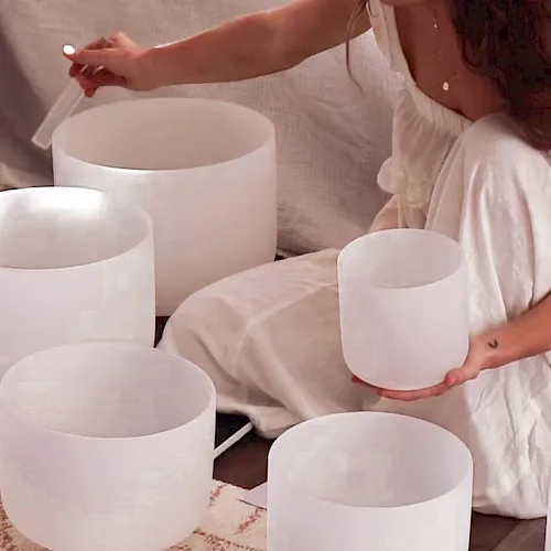 A woman in a white dress holds a white candle bowl while others sit on a table, creating a calm, minimalist vibe.