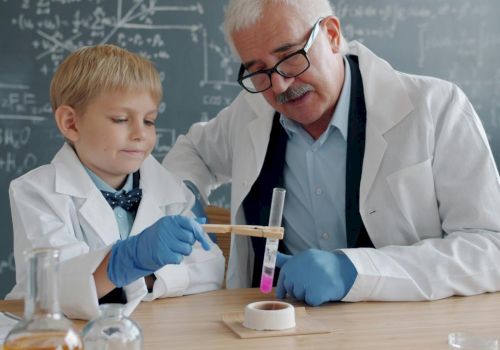 A child in a lab coat and gloves helps an older scientist with test tubes and chemicals in a classroom lab.